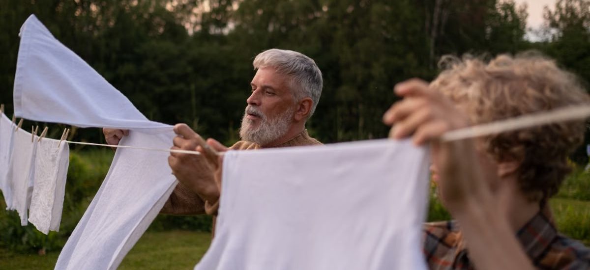 Elderly man and teenage boy working together, hanging laundry in a green outdoor setting.